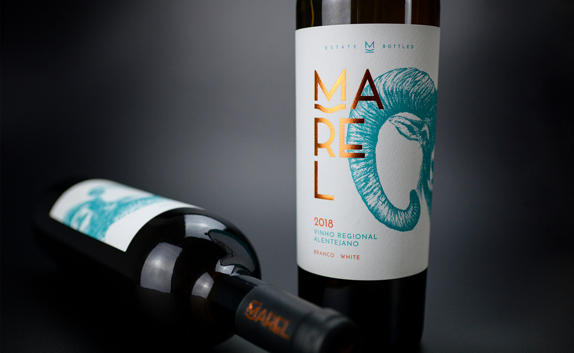 Duas garrafas de vinho Marel Branco. Uma com o rótulo bem visível: o chifre do Marel é azul-turquesa e lê-se: MAREL, 2018 - Vinho Regional Alentejano.
