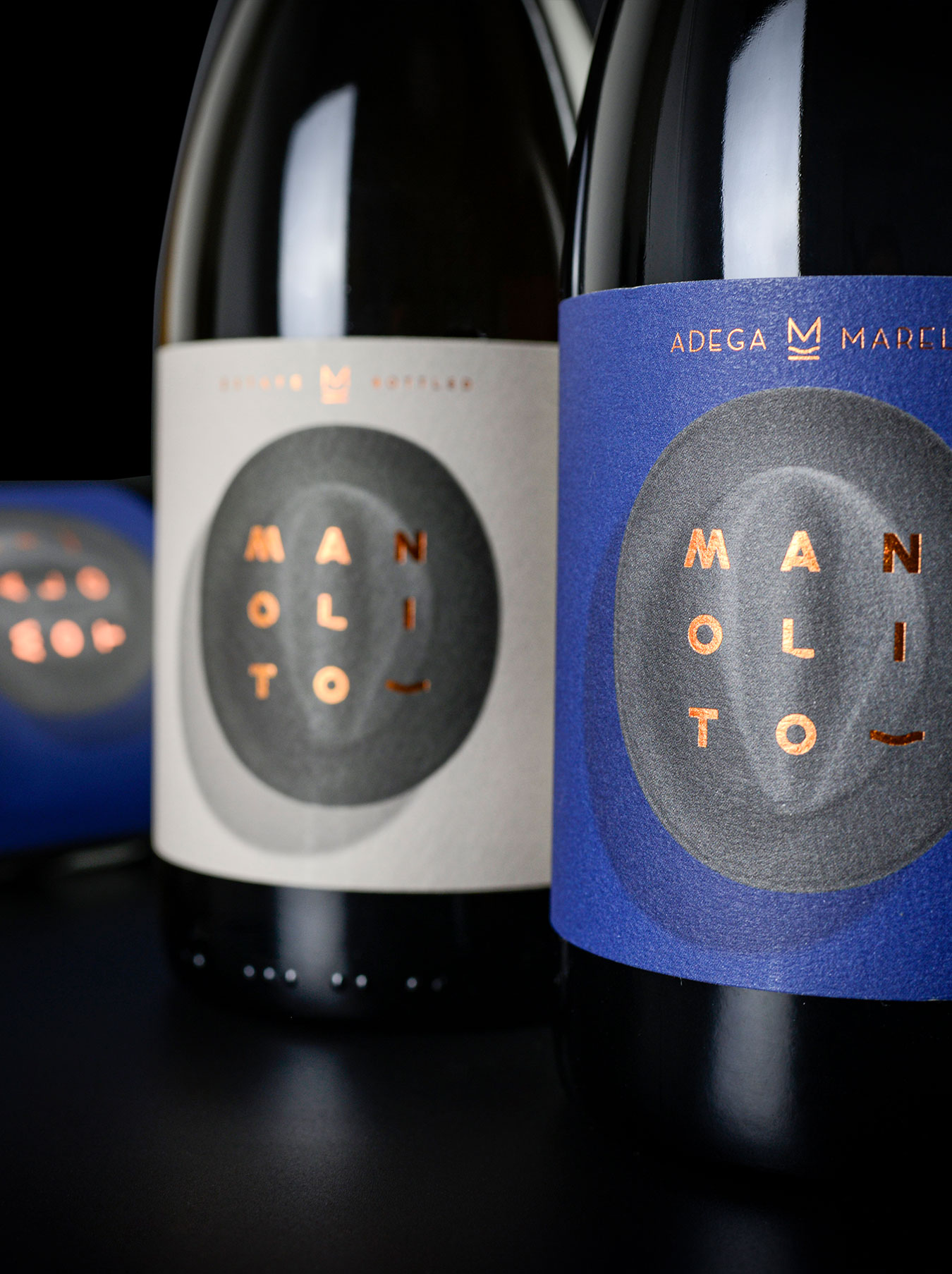 Três garrafas de vinho Manolito da Adega Marel: duas de vinho tinto e uma de vinho branco. Os rótulos são azuis no tinto e creme no branco, com imagens de chapéus fedora aveludados.